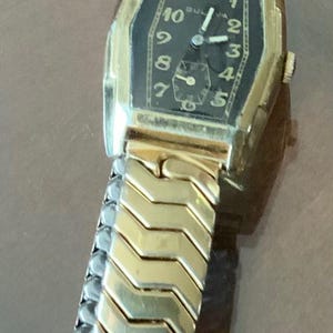 May include: A gold-toned wristwatch with a black face and a gold metal band. The watch face has Roman numeral hour markers and a small second hand.