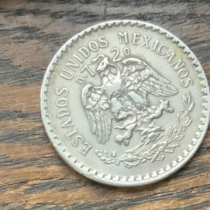 May include: A silver coin featuring the Mexican coat of arms, an eagle with a snake in its beak. The coin is inscribed with "ESTADOS UNIDOS MEXICANOS" and the year "20". The coin has a ridged edge.