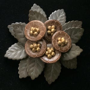 May include: A floral brooch featuring five brown flowers with gold-colored centers, surrounded by silver-colored leaves. The brooch is likely made of metal and has a detailed, textured design. The overall shape is circular, resembling a stylized flower arrangement.