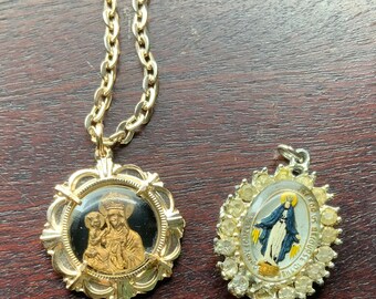 May include: Two gold tone pendants on chains. One pendant features a round, black and gold image of the Virgin Mary and baby Jesus. The other pendant is oval shaped with a clear glass center and a gold tone border with rhinestones. The text on the oval pendant reads "Our Lady of Lourdes".