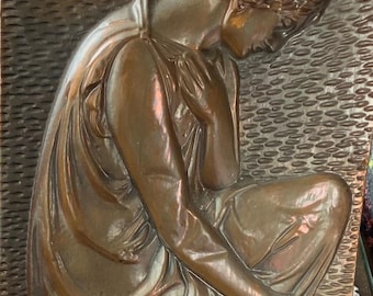 May include: A copper wall plaque featuring a relief sculpture of a woman in a flowing robe, kneeling with her head bowed. The woman's hair is styled in a bun and she is holding a small object in her hand.