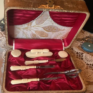 May include: A vintage vanity set in a brown leatherette case with a red satin lining. The set includes a nail file, cuticle pusher, scissors, and a small brush. The case is decorated with gold accents.