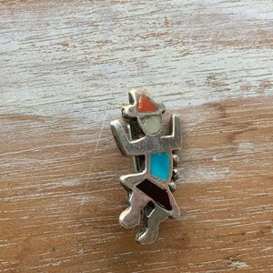 May include: A silver charm with a stylized figure in turquoise, red, black, and white stones.