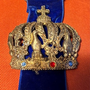 May include: A vintage gold-toned metal badge with a crown design, featuring a blue ribbon. The badge has a cross on top and is adorned with red and blue jewels. The text "6. IN WANDERTAGE TV FORST" is visible.