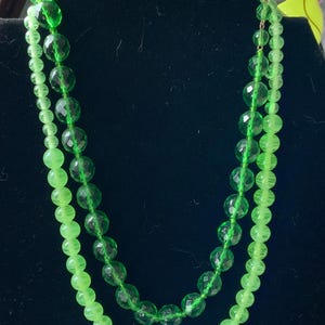 May include: A double strand necklace made of green glass beads. The beads are different sizes and shapes, with some being round and others being faceted. The necklace is displayed on a black velvet background.