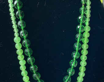 May include: A double strand necklace made of green glass beads. The beads are different sizes and shapes, with some being round and others being faceted. The necklace is displayed on a black velvet background.