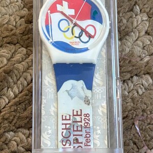 May include: A white Swatch watch with a blue, red, and white band. The watch face features the Olympic rings and the Swiss flag. The watch is in a clear plastic case with the words "Swatch Quartz" and "Swiss Made" printed on it. The case also has a sticker with the Olympic flame and the words "Atlanta 1996".