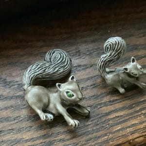 May include: A pair of silver squirrel earrings with green eyes. The squirrels are detailed with a textured fur pattern.