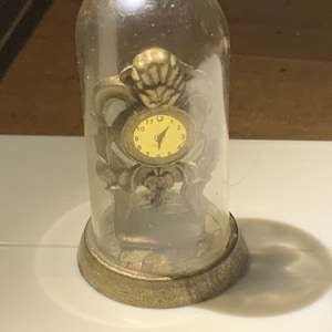 May include: A miniature clock encased in a clear glass dome. The clock has a cream-colored face with black numerals and hands. The clock is set in an ornate, gold-colored metal frame. The base of the dome is also gold-colored.