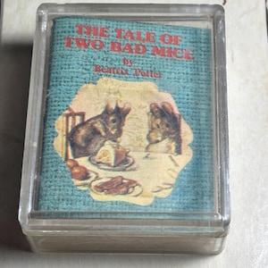May include: A small, clear plastic case containing a miniature book. The book cover features the title "THE TALE OF TWO BAD MICE" by Beatrix Potter, with an illustration of two mice at a table with food. The background is a light blue.