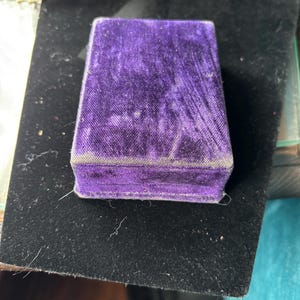 May include: A small, rectangular box covered in vibrant purple velvet. The box has a hinged lid and a soft, textured appearance. The box is sitting on a black surface, with a partially visible burgundy velvet box in the background.