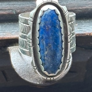 May include: A silver ring with an oval, deep blue stone. The stone is set in a scalloped bezel. The ring band has a layered design with etched details. The ring is likely a piece of jewelry.
