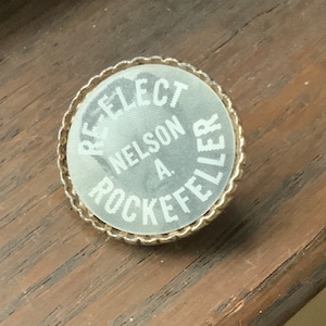 May include: A white button with a gold rim. The button says "Re-Elect Nelson A. Rockefeller".