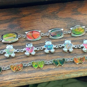 May include: Three silver bracelets with colorful, iridescent, oval-shaped charms. The charms are arranged in a pattern of bears, hearts, and abstract designs.