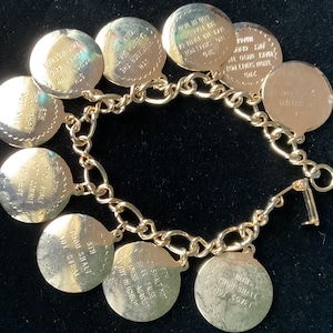 May include: Gold-toned bracelet featuring circular charms with engraved text. The charms are connected by a chain link design and fastened with a clasp. The text on the charms is partially visible.