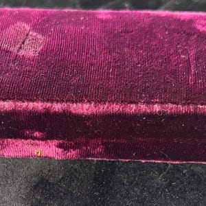 May include: A rectangular, burgundy velvet box with a hinged lid. The box has a plush, textured surface and a slightly faded appearance. The box is likely used for jewelry or other small valuables.