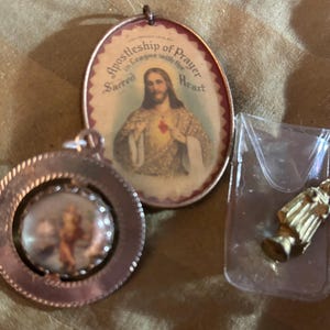 May include: Three religious pendants and charms. One oval pendant features an image of Jesus with the text "Apostleship of Prayer". A round pendant with a cherub image and a small gold statue in a plastic sleeve.