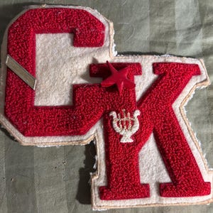 May include: A varsity-style patch featuring the letters CK in red chenille embroidery on a cream felt background. The patch includes a small red star, a metallic silver bar accent, and a white embroidered lyre symbol centered on the letter K.
