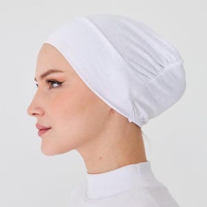 Può includere: Un copricapo bianco, probabilmente un berretto o un cappuccio, è mostrato di profilo. Il tessuto sembra morbido e leggero, con un dettaglio arricciato sul retro. Il copricapo è indossato sulla testa.