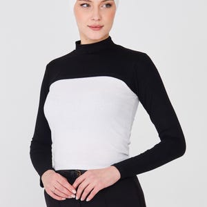May include: A black and white long-sleeved top with a high neckline. The top has a white panel across the chest and black sleeves. A white head covering is also visible. The model is wearing black pants.