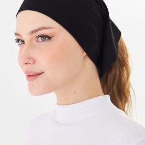 May include: A black headscarf, likely made of a soft fabric, is worn on the head. The head covering is neatly styled, with a portion of the hair pulled back into a ponytail. The person is wearing a white turtleneck.