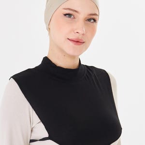 May include: A beige head covering and a black modesty panel are worn over a cream-colored long-sleeved top. The modesty panel has a high neckline and covers the chest area. The background is plain white.