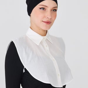Puede incluir: Un gorro negro y un cuello desmontable blanco con botones. El cuello se lleva sobre una camiseta negra de manga larga. La imagen está sobre un fondo blanco.