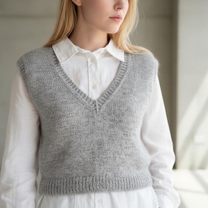 Pode incluir: Um colete cinza claro com decote em V. O colete é feito de material de malha e usado sobre uma camisa branca com gola. Possui bainha canelada e um design simples. A modelo está usando o colete.