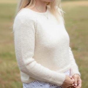 Könnte beinhalten: Cremefarbener Strickpullover mit langen Ärmeln. Der Pullover hat einen Rundhalsausschnitt und ein leicht strukturiertes Aussehen. Die Ärmel sind eingesetzt, und der Gesamtstil ist lässig und bequem. Der Pullover ist aus weichem Garn gefertigt.