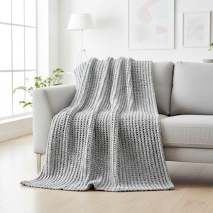 May include: A grey knitted throw blanket draped over a light grey sofa. The blanket has a textured, waffle-like pattern. The sofa is in a well-lit room with a window and plants.