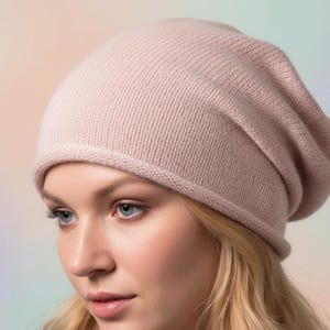 May include: A soft, light pink beanie hat. The hat is made of a knitted material and has a ribbed brim. The beanie is worn by a person with blonde hair and blue eyes. The background is a soft gradient of colors.