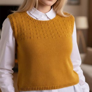 以下が含まれることがあります： マスタードイエローのニットベスト。クルーネックで、胸上部にテクスチャードダイヤモンド柄。白い襟付きシャツの上に着用。全体的にカジュアルで快適なスタイルで、重ね着に適しています。