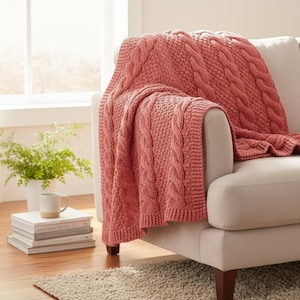 May include: A coral-colored knitted throw blanket with a cable knit design draped over the arm of a beige sofa. The blanket has a textured appearance and adds a cozy element to the living room setting. A stack of books and a mug are visible.