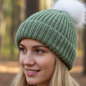 May include: A green knit beanie with a white faux fur pom-pom. The hat is ribbed and has a folded brim. The background is a blurred forest scene.