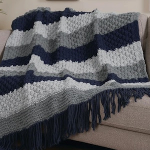 May include: A navy blue, grey, and light grey knitted throw blanket with fringe. The blanket is draped over a beige sofa. The textured pattern creates a cosy, inviting aesthetic. The fringe is navy blue.
