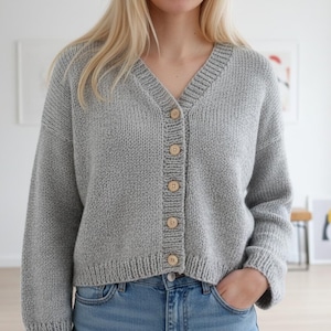 以下が含まれることがあります： Vネックと長袖のライトグレーのニットカーディガン。セーターは、5つの木製ボタンが付いたボタンアップフロントです。モデルはブルージーンズを着用しています。カーディガンは快適でカジュアルなスタイルです。