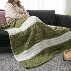 May include: A green and white striped crochet blanket draped over a black sofa. The blanket has a textured appearance and is folded over a person sitting on the sofa. A pink pillow rests on the sofa.