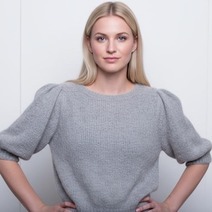 Può includere: Un maglione grigio chiaro con scollo tondo e maniche a sbuffo. Il maglione ha una trama a coste ed è realizzato in un morbido materiale a maglia. La modella indossa il maglione e ha le mani sui fianchi.