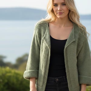 Könnte beinhalten: Ein salbeigrüner Strickcardigan mit hochgekrempelten Ärmeln. Der offene Pullover hat einen Kragen und wird über einem schwarzen Oberteil und dunklen Jeans getragen. Der Hintergrund zeigt eine verschwommene Ansicht von Wasser und Grün.