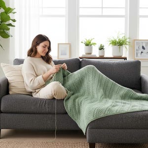 Könnte beinhalten: Eine Frau strickt eine hellgrüne Decke auf einem grauen Ecksofa. Die Decke hat ein strukturiertes Muster. Der Raum hat ein großes Fenster, Pflanzen und gerahmte Kunstwerke. Die Frau trägt einen beigen Pullover und eine Hose.