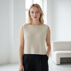 Peut inclure: Une femme porte un haut sans manches en tricot crème et un pantalon noir. Le haut a une texture côtelée et une encolure arrondie. Elle se tient dans une pièce lumineuse et minimaliste avec une chaise blanche.