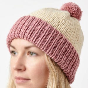 May include: A knit beanie featuring a cream-colored crown and a wide, ribbed cuff in a dusty rose pink. The hat is topped with a matching dusty rose pom-pom. The construction shows a clear contrast between the smooth knit top and the textured ribbing.