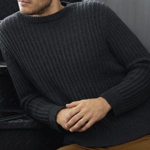 Puede incluir: Un suéter gris oscuro de cuello redondo con un patrón de punto texturizado. El suéter de manga larga lo lleva una persona, con los brazos apoyados en el regazo. El suéter está hecho de un material grueso y cálido, adecuado para climas más fríos.