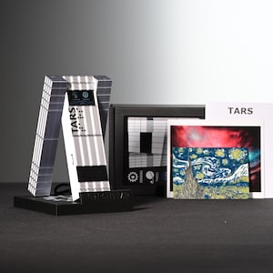 Può includere: Un'esposizione con un modello TARS a strisce bianche e grigie con la scritta "TARS", una scatola nera con una grafica e una stampa de "La notte stellata" di Van Gogh. L'esposizione è su una superficie nera.