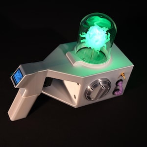 May include: A futuristic, silver-colored ray gun prop with a glowing green orb encased in a glass dome. The gun features a small blue screen, a metallic dial, and a purple tube detail. The overall design is sleek and modern.