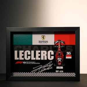 Puede incluir: Obra de arte enmarcada con un modelo de coche de carreras Ferrari de Fórmula 1 rojo. La obra incluye el nombre "CHARLES LECLERC", el número "16" y una firma. El fondo tiene el logotipo de Ferrari y los colores de la bandera italiana.