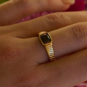Puede incluir: Un anillo de oro con una piedra preciosa oscura rectangular. El anillo tiene una banda texturizada con un diseño acanalado. La piedra preciosa está engastada en un bisel de oro. El anillo está en un dedo.