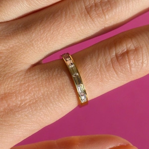 May include: A gold band ring with rectangular, clear gemstones set across the top. The ring is on a finger, with a bright pink background.