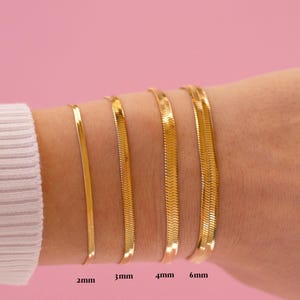 Op de afbeelding: Een collectie gouden visgraatarmbanden in verschillende breedtes, van 2 mm tot 6 mm, getoond op een pols. De armbanden zijn gerangschikt in volgorde van toenemende breedte, met de afmetingen duidelijk aangegeven tegen een roze achtergrond.