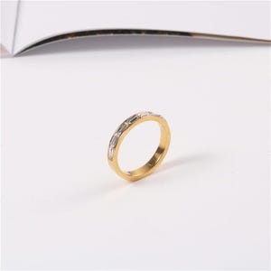 May include: A gold-colored ring with rectangular, clear gemstones set around the band. The ring is on a white surface, with a blurred background.